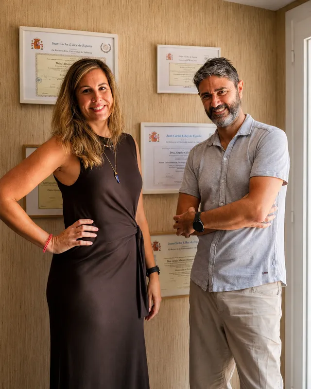 Ángela y Jesús de pie junto a la pared con sus títulos y diplomas universitarios enmarcados